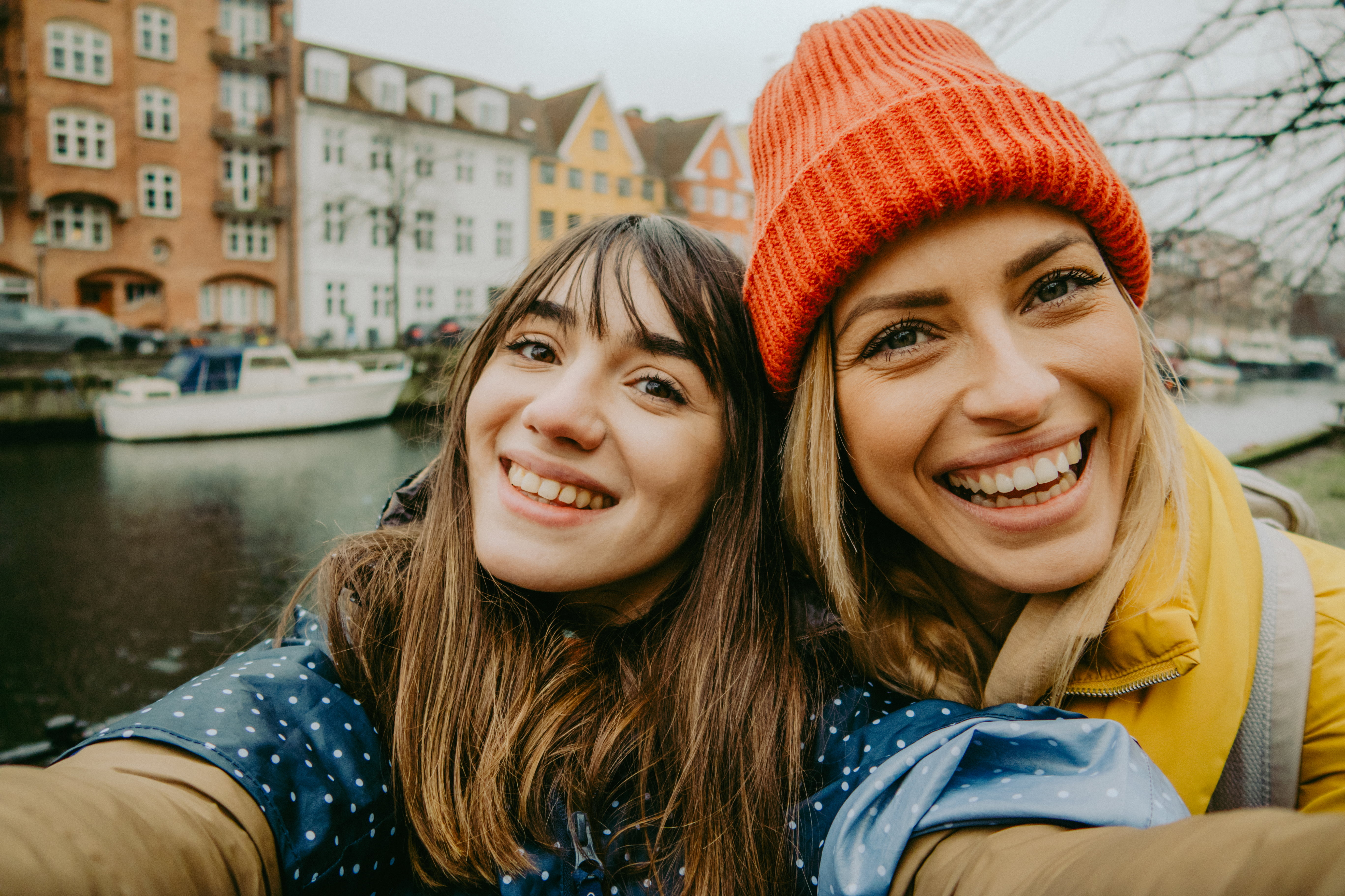 To smilende venninner tar en selfie med en pittoresk kanal og fargerike bygninger i bakgrunnen, som representerer glede og fellesskap i hverdagsøyeblikkene.