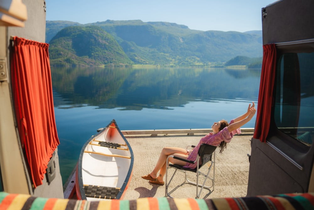 Kvinne som sitter i solstol ved vannet, med kano ved brygga. Strekker armene i været og nyter sommersolen.