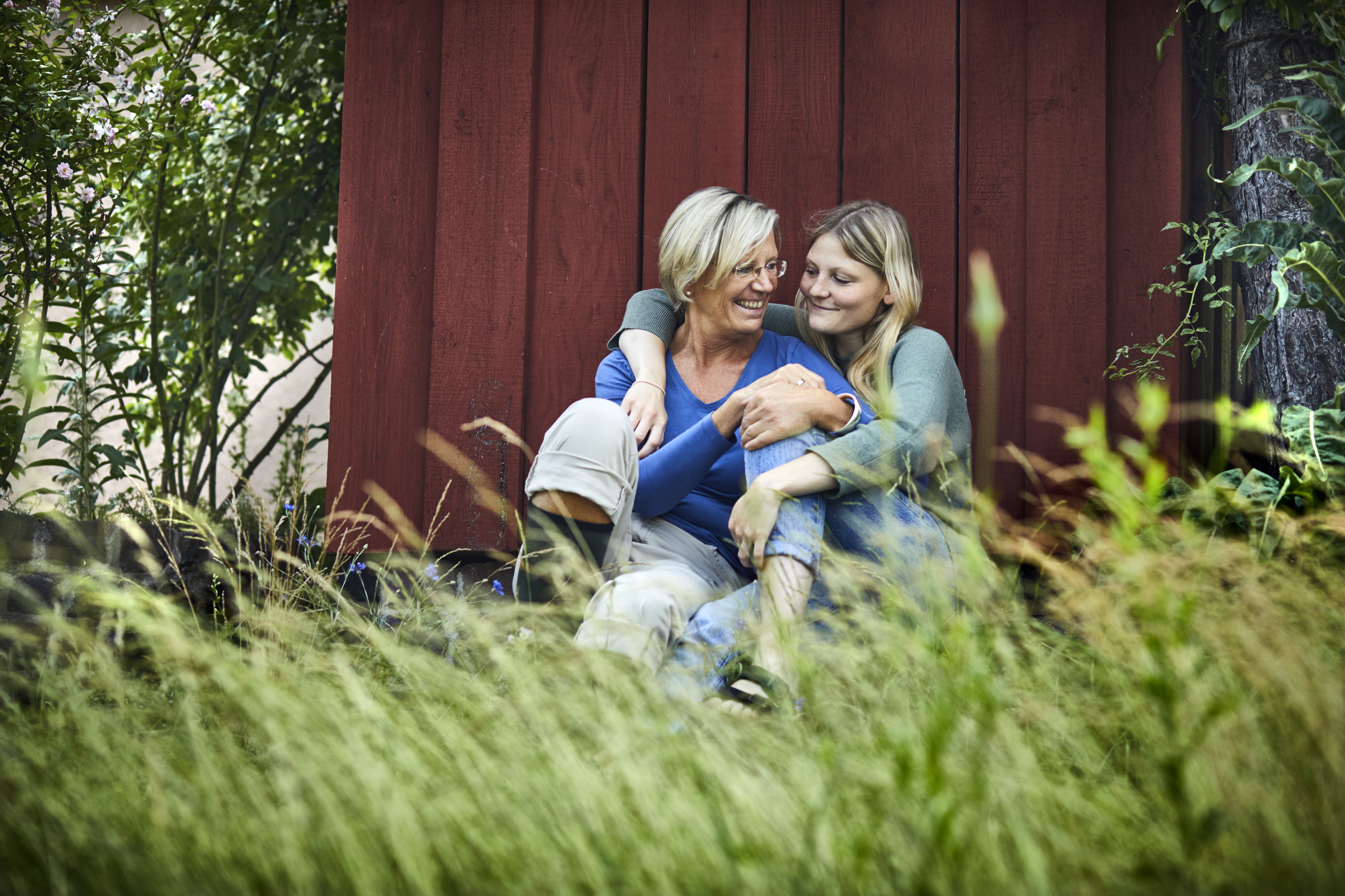 Mor og datter som sitter i gresset utenfor rød hytte